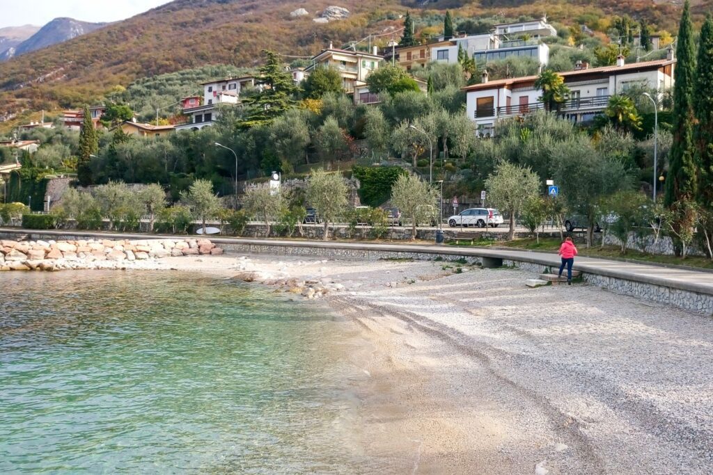 A person in a pink jacket sits on a stone wall by a pebbly beach with clear water. Houses and greenery cover the hillside in the background under a partly cloudy sky.