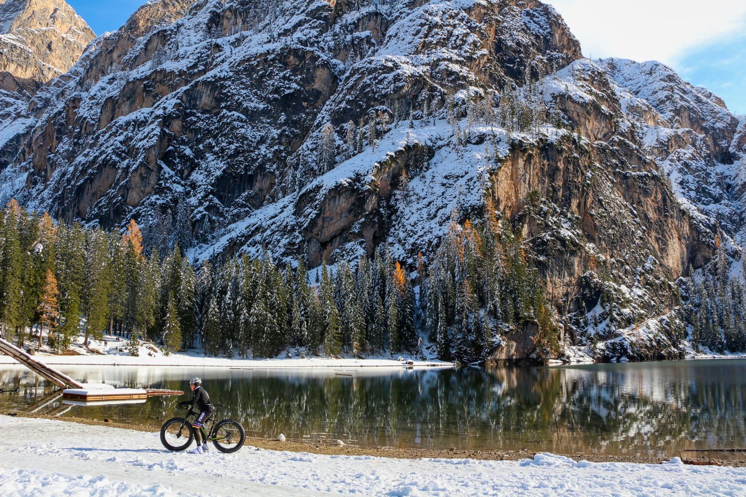 A person rides a bicycle on snow beside a calm lake, with tall evergreen trees and steep, rocky mountains covered in snow in the background under a partly cloudy sky.