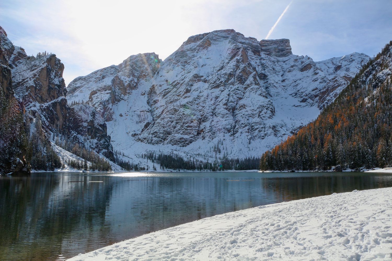 Snow-covered mountains rise above a calm lake, reflecting their snowy peaks. Pine trees with patches of autumn colors line the water’s edge, and the foreground shows snow on the shore under a bright, partly cloudy sky.
