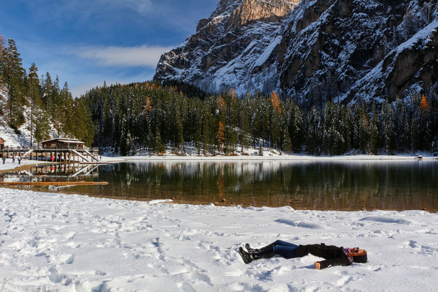 A person lies on their back in the snow beside a calm, partially frozen lake surrounded by pine trees and snowy mountains under a blue sky. A wooden dock and building are visible on the left.