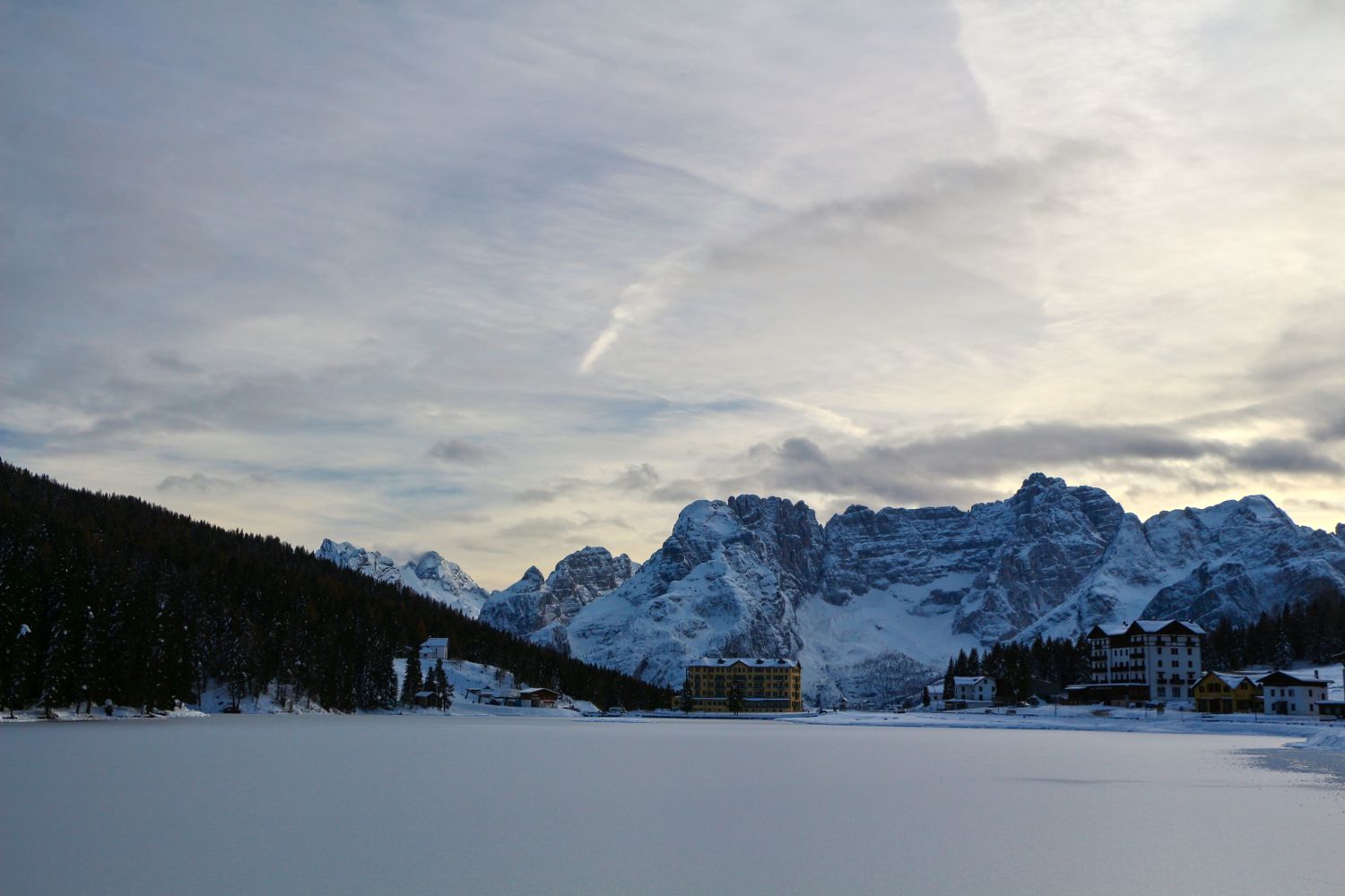 Snow-covered mountains under a cloudy sky, with buildings nestled at the base and a frozen lake in the foreground, surrounded by trees on one side.