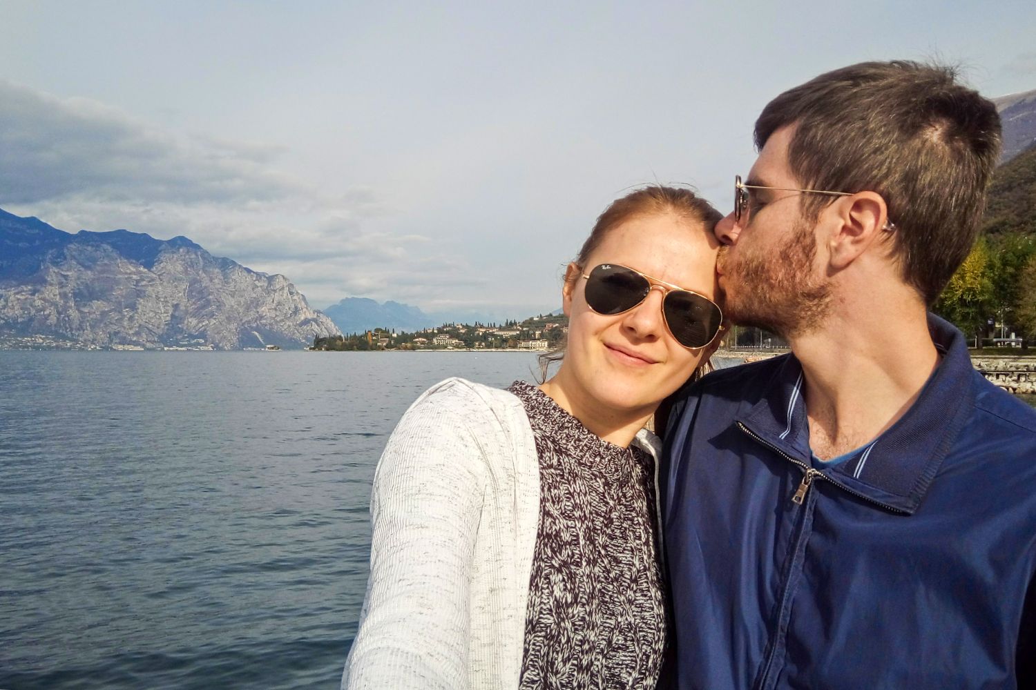 About 1 A couple wearing sunglasses takes a selfie by a lake, with mountains and a small town in the background. The man kisses the woman on the forehead as they both smile, enjoying a sunny day outdoors.