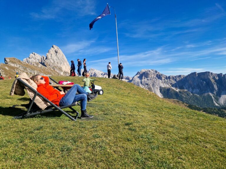 A person relaxes on a lounge chair on a grassy mountain slope, while others walk nearby. A flag waves on a tall pole. Rocky mountains and a blue sky are in the background.