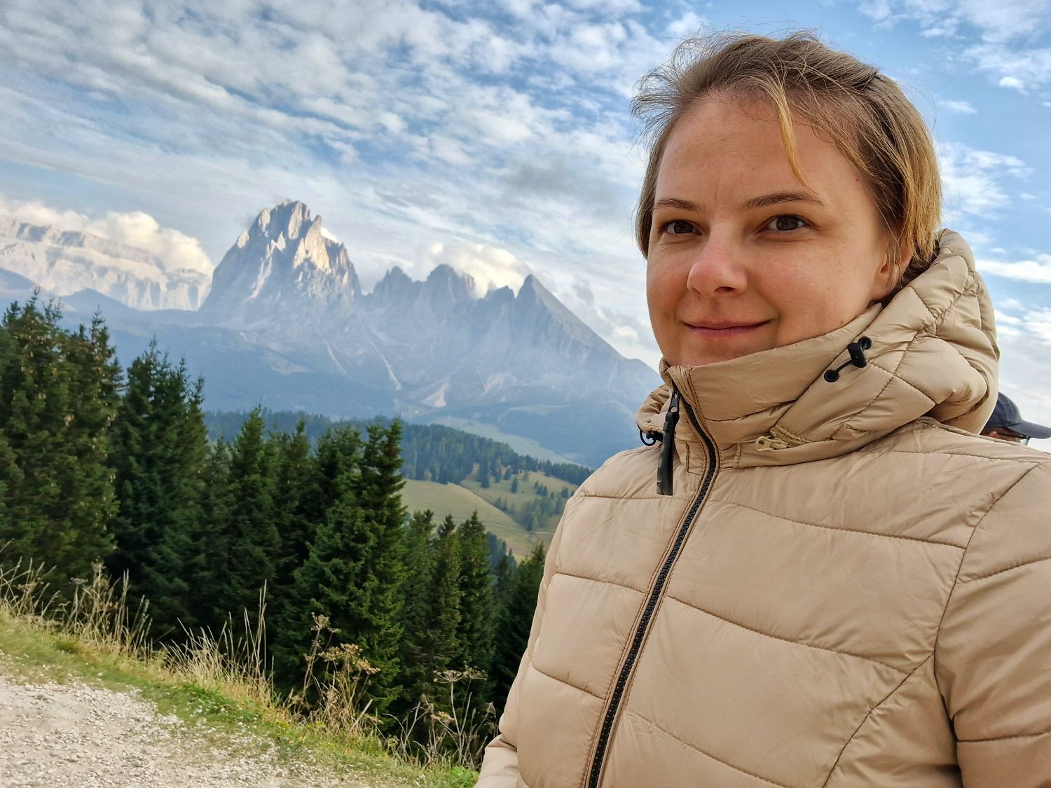 Home 10 A woman in a beige puffer jacket stands outdoors, smiling slightly, with green pine trees and rugged, snow-dusted mountains in the background under a partly cloudy sky.