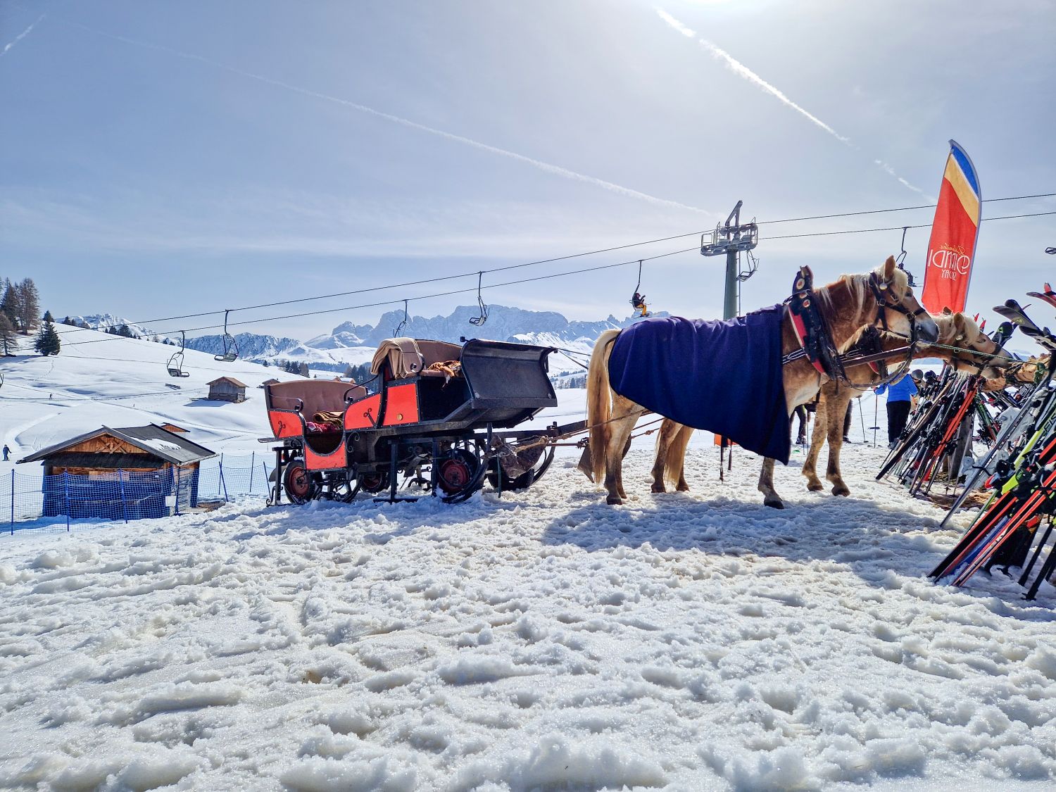 Two horses wearing harnesses and blankets stand in the snow beside a red carriage near a ski area. Skis are lined up on racks to the right while chairlifts, small wooden huts, and distant mountains sit under a bright sunny sky.