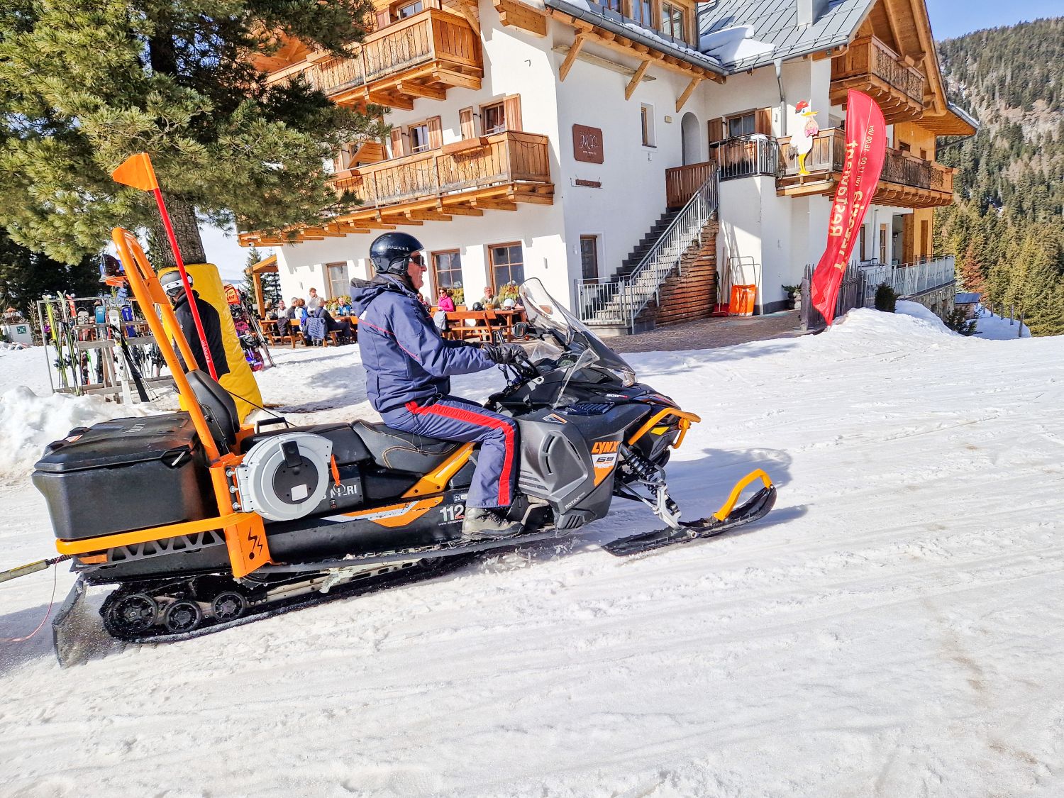 Person in a helmet drives an orange snowmobile across a snowy path beside a wooden alpine lodge with balconies. Ski racks, outdoor seating with people, and a red flag sign are visible near the building with forested slopes rising behind it.