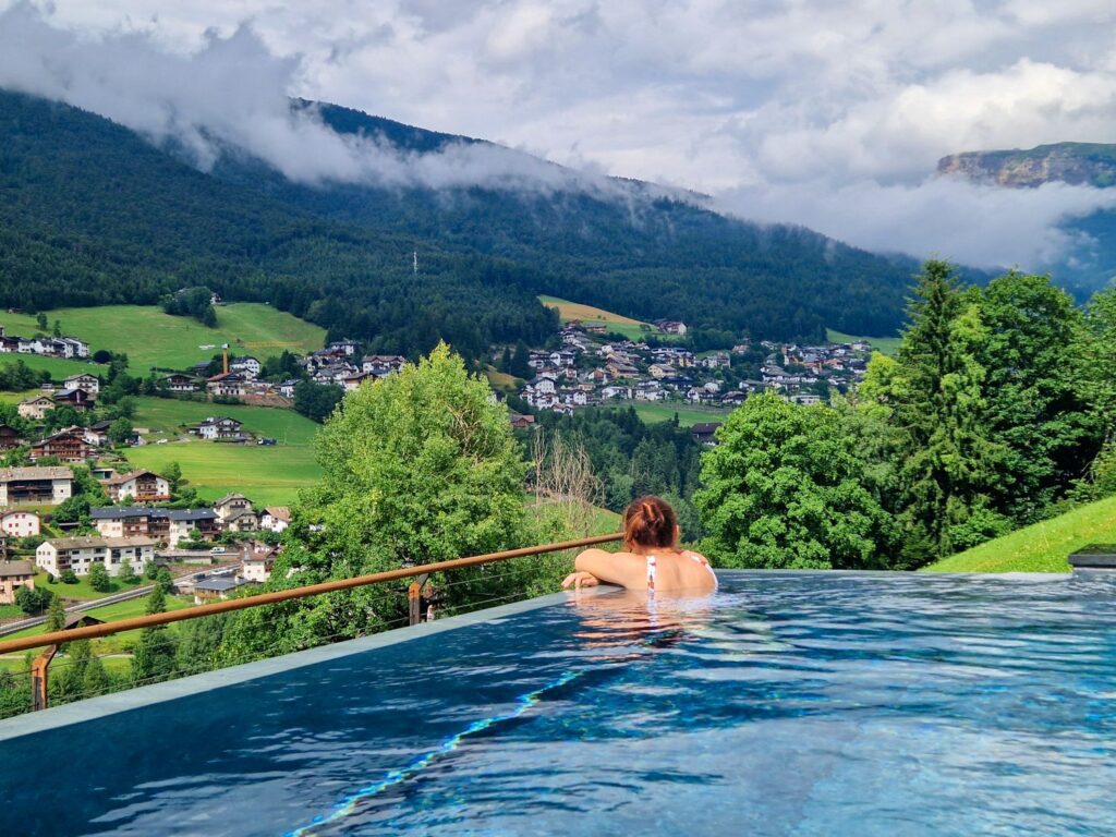 A person relaxes at the edge of an infinity pool overlooking a scenic valley with green hills, trees, and a village. Misty mountains and clouds are visible in the background.