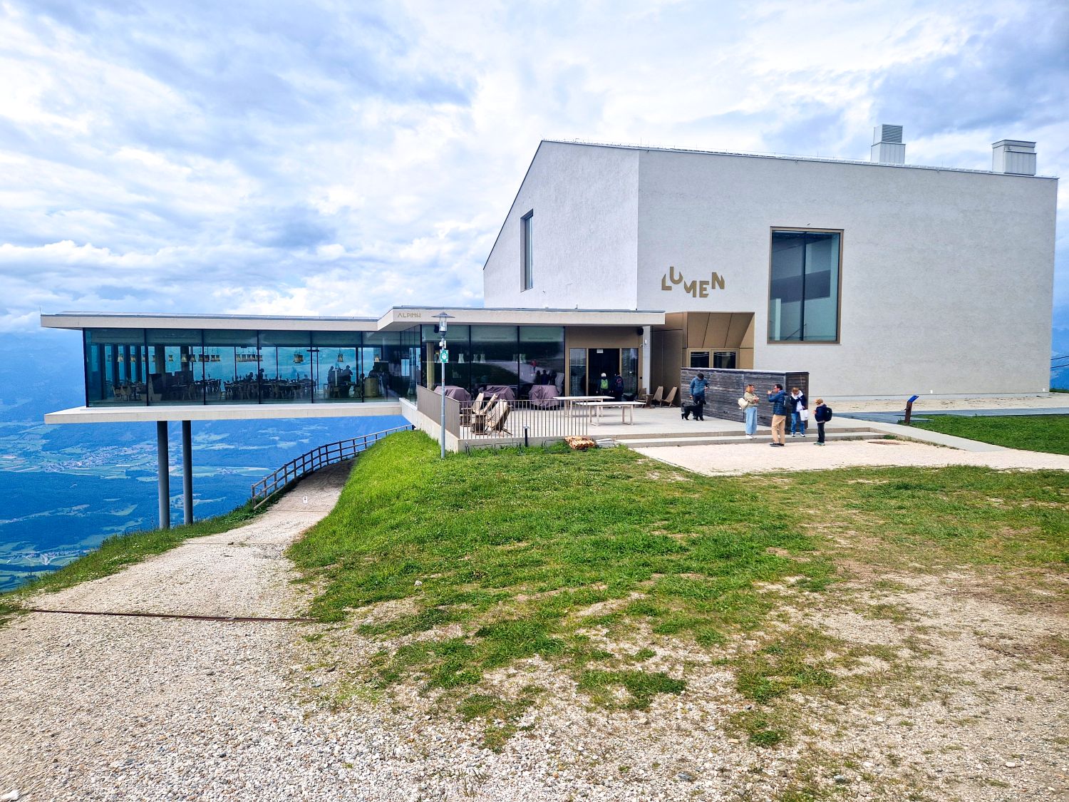 A modern white building labeled "LUMEN" sits on a grassy hilltop, with large glass windows overlooking a scenic mountain landscape. Several people stand outside under a cloudy sky.