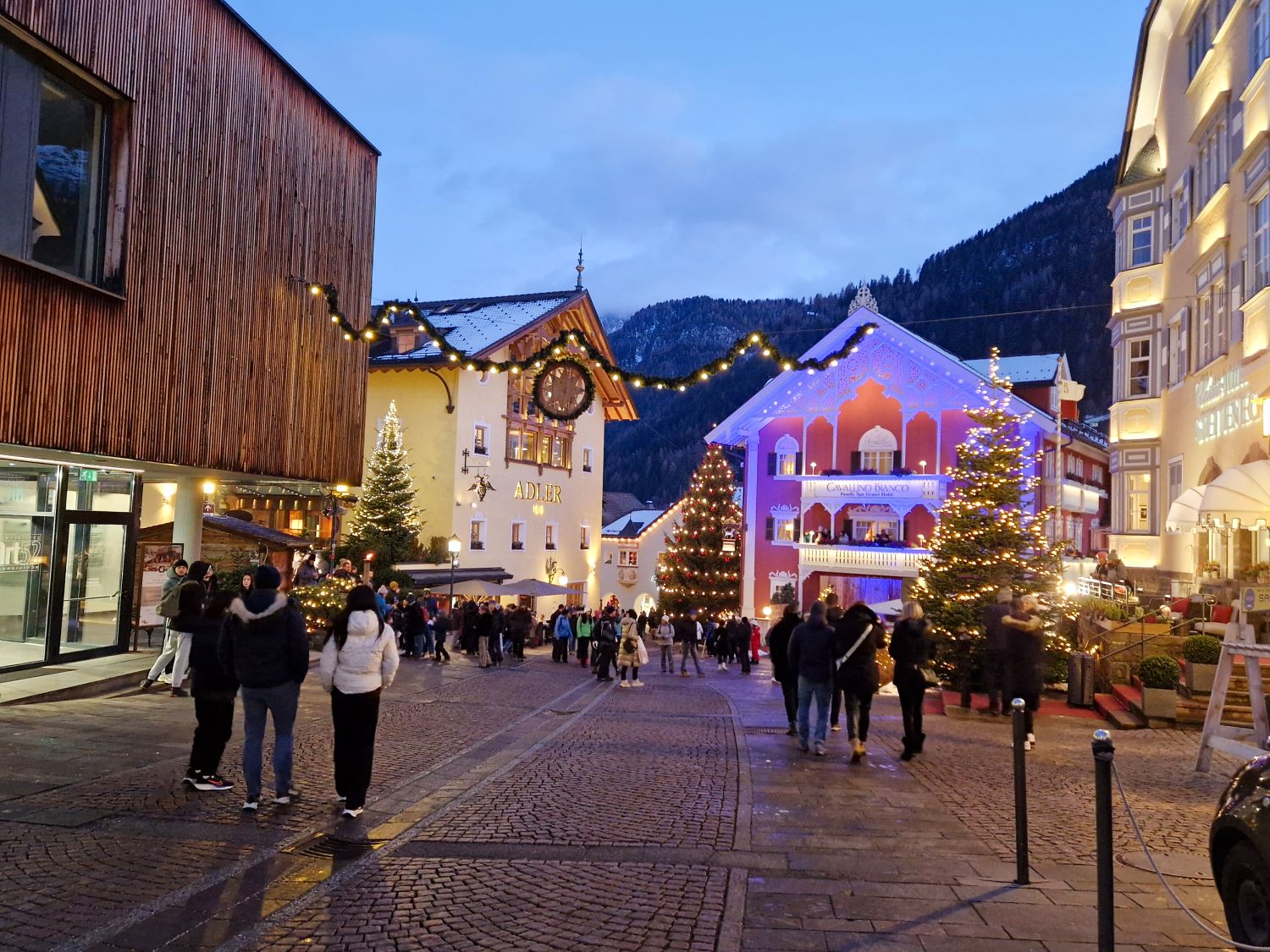 Festive town square at dusk with people walking along a cobblestone street in Ortisei, Val Gardena lined with lit Christmas trees and glowing string lights overhead. Hotel buildings decorated with warm lights include one with a clock and the word "ADLER" and another reading "CAVALINO BANCO" set against dark mountain silhouettes in the background.