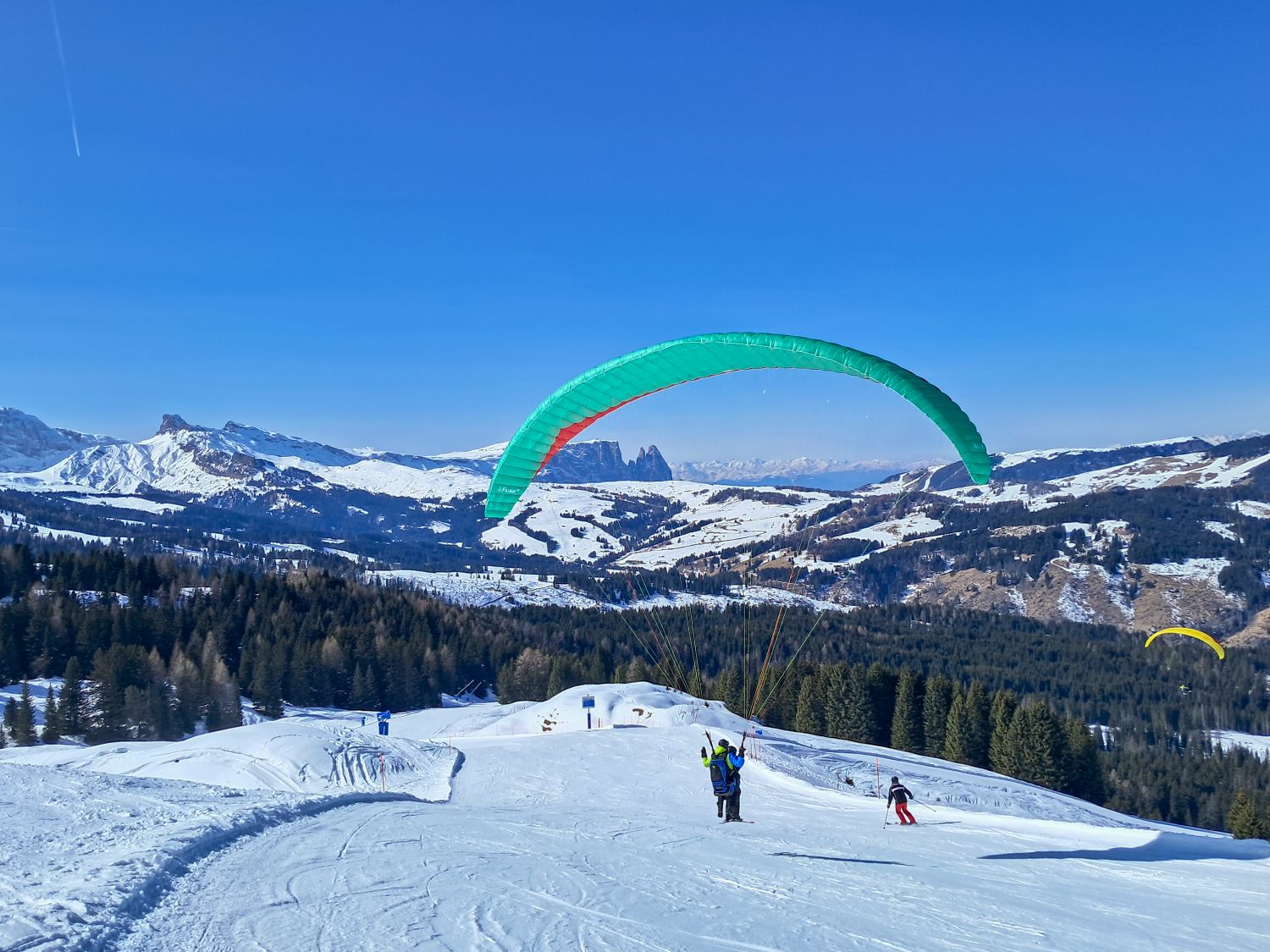 a green paraglider prepared to launch on a snowy ridge in Monte Pana with wide alpine views of rolling hills and jagged peaks in the distance. Another skier moves downhill nearby while a second paraglider floats over the forested valley under a clear blue sky.