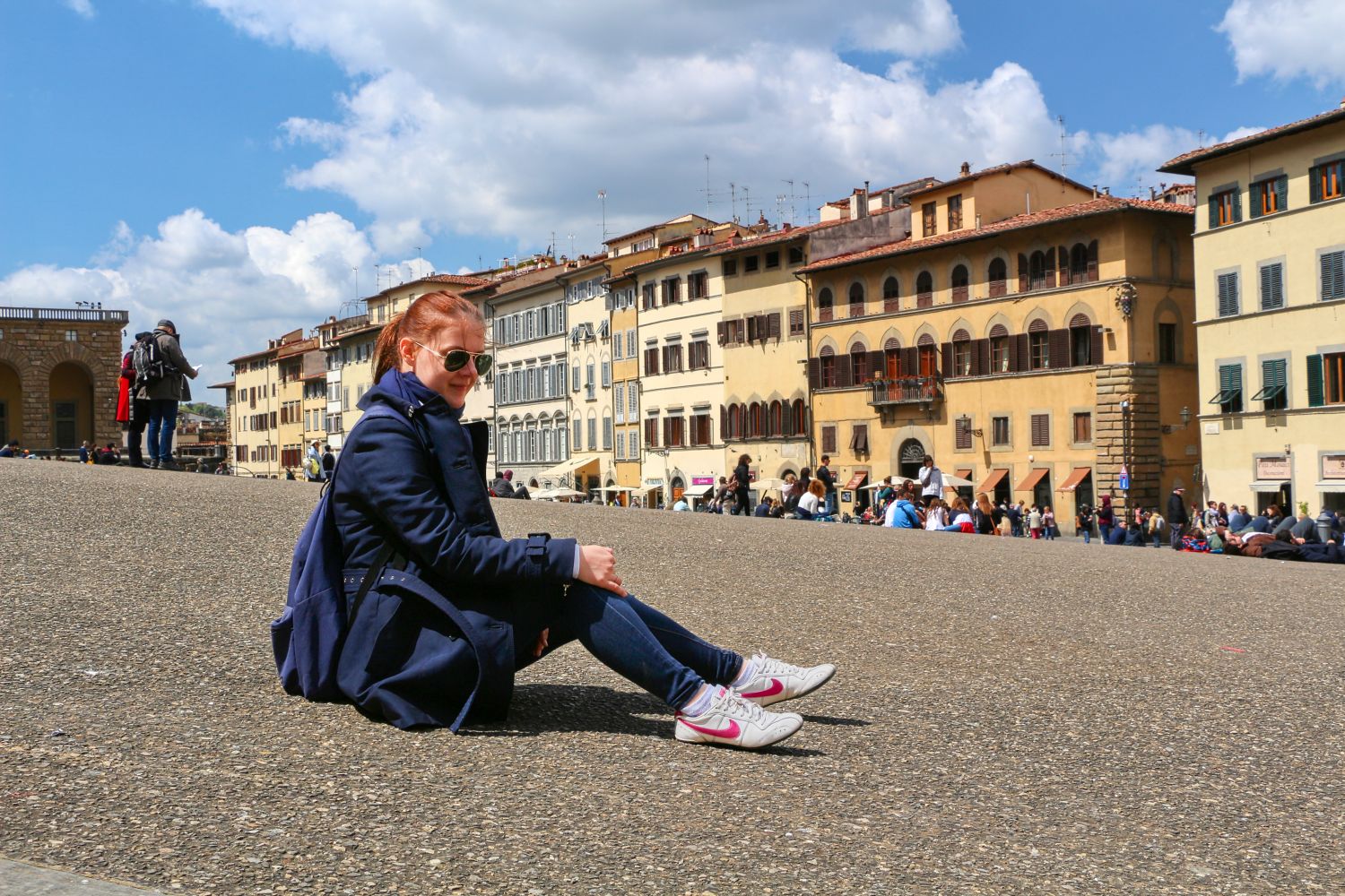 A person in a navy coat, jeans, and white sneakers sits on a stone plaza with historic yellow buildings and many people in the background under a partly cloudy sky.