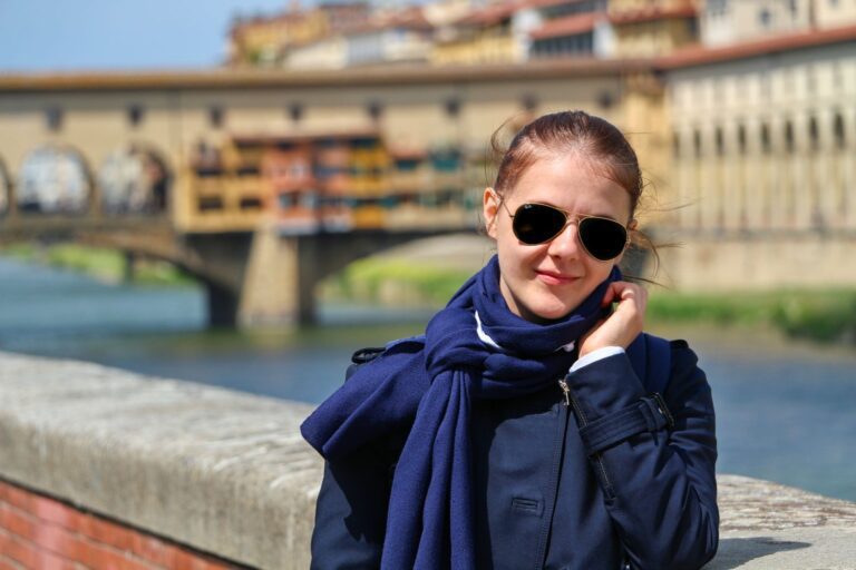A woman wearing sunglasses, a navy coat, and a scarf stands by a stone railing with a blurred view of the Ponte Vecchio bridge in Florence, Italy, and a river in the background.
