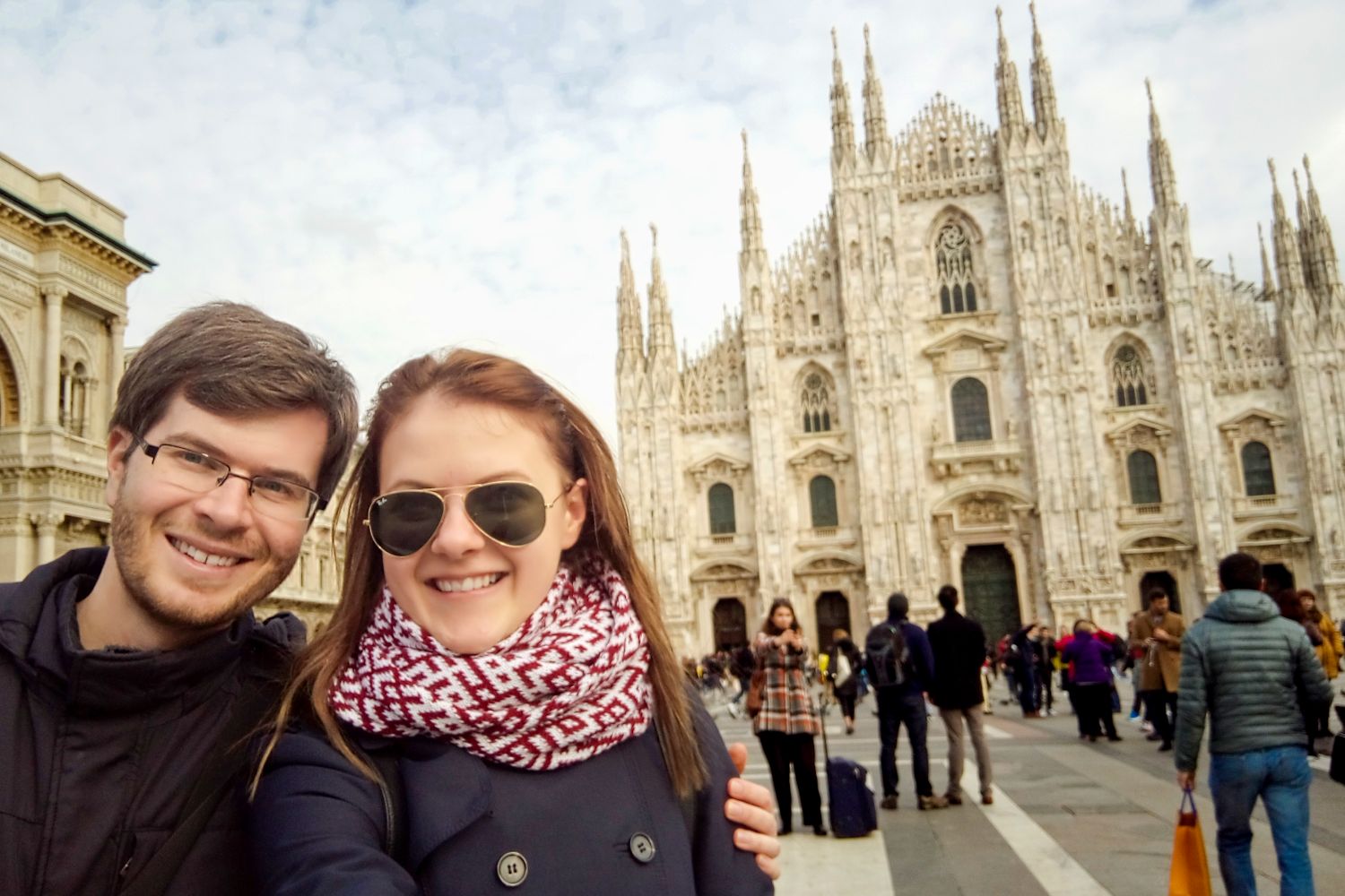 A smiling couple takes a selfie in front of the Milan Cathedral in Italy. The ornate, Gothic-style facade of the cathedral is visible, with people walking and gathering in the plaza.