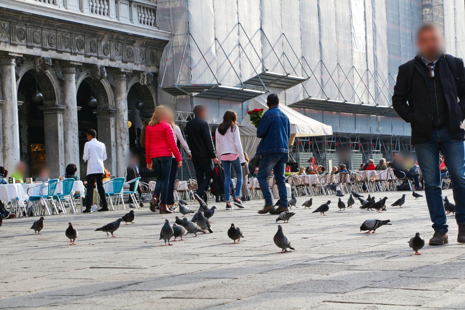 People walk and stand in an open square with pigeons on the ground. Outdoor café seating with blue chairs is visible to the left, and a large building is under construction in the background. Faces are blurred for privacy.