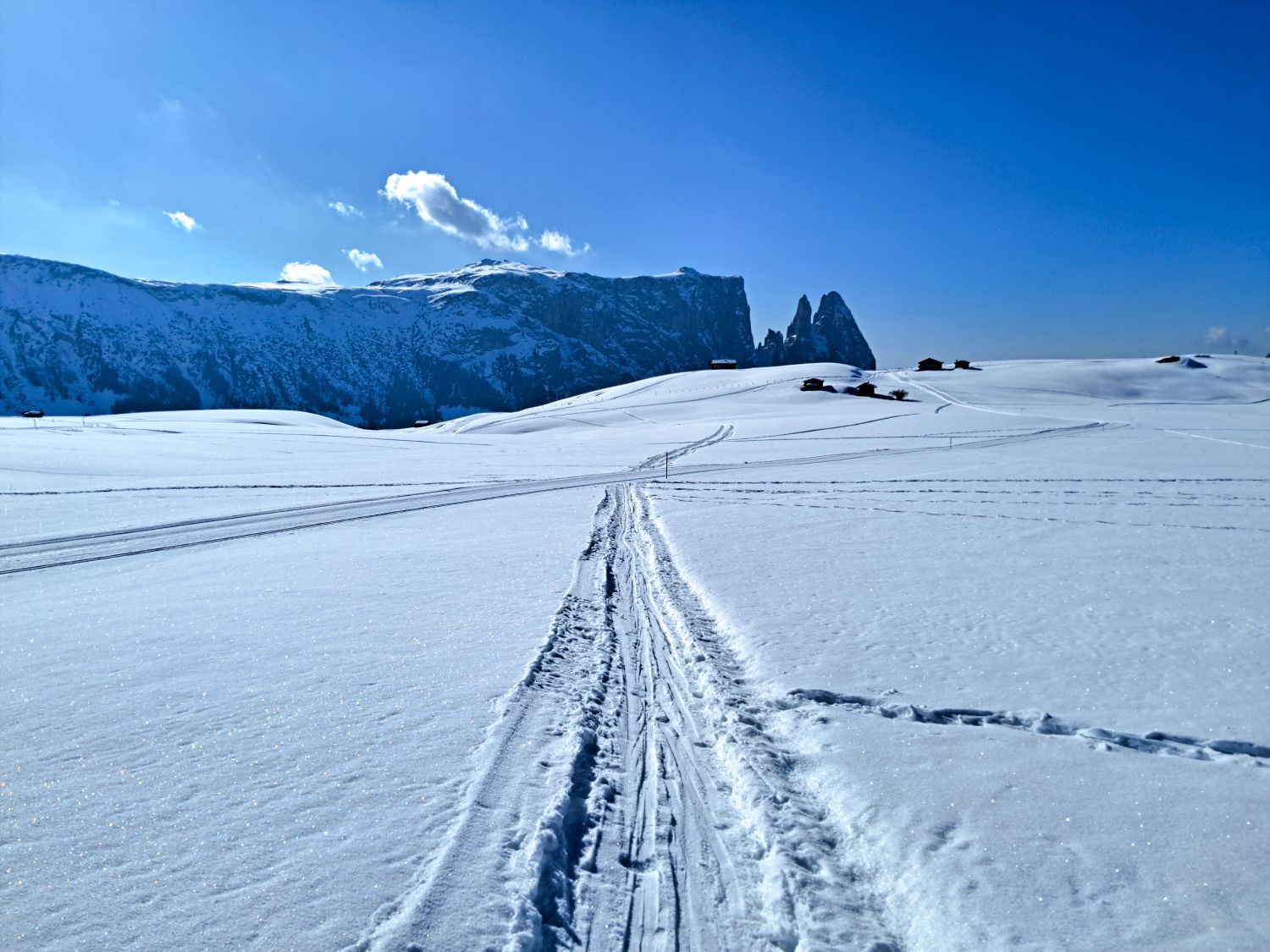 Snow covered landscape with narrow ski tracks leading into the distance toward a range of rugged mountains under a bright blue sky. A few small wooden huts sit scattered on the rolling hills, emphasizing the quiet and remote winter setting.