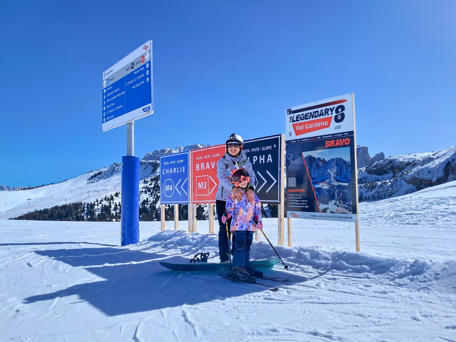 Adult and child in ski gear stand on a snowy slope in front of trail signs while smiling at the camera. Signs behind them read "CHARLIE M4" "BRAVO M3" and "ALPHA" with directional arrows and a larger board that says "THE LEGENDARY 8 Val Gardena" and "BRAVO". Snow covered mountains and a clear blue sky stretch across the background.