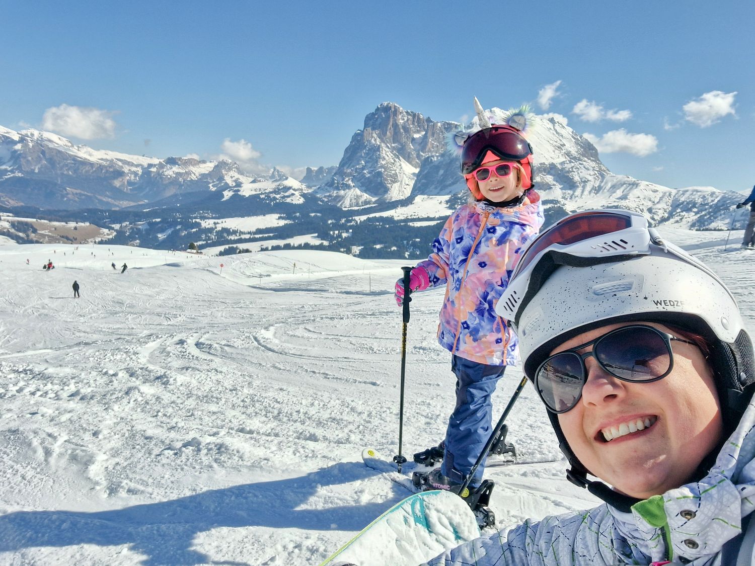 Smiling adult and child in ski gear pause on a snowy slope with mountains behind them on a sunny day. The child in a colorful jacket and pink sunglasses stands on skis while the adult takes a close up selfie, capturing a playful moment during a day of skiing. Kristine and Emma on the Paradiso slope skiing in Alpe di Siusi