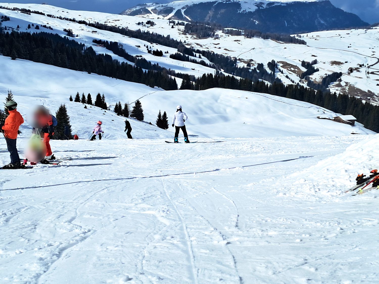 Several people skiing and snowboarding on a wide snowy slope with rolling hills and pine trees in the background. A person in a white jacket stands facing downhill while others gather to the side, with scattered cabins and winding trails visible across the mountain landscape.