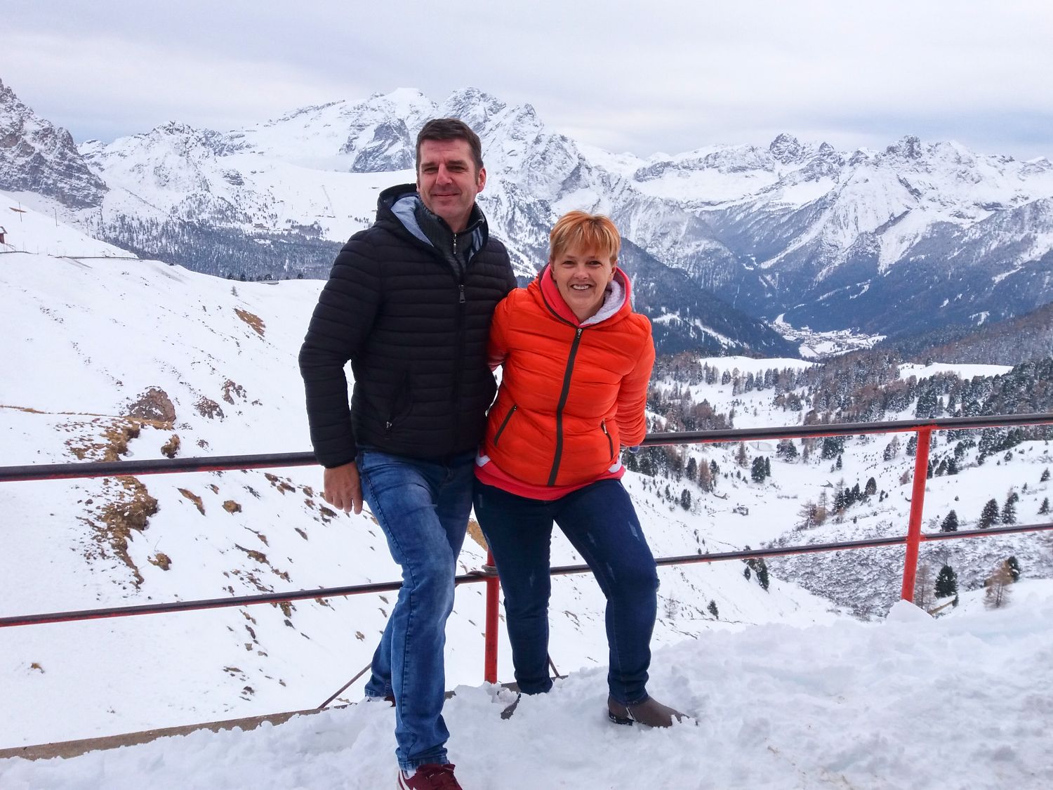 Home 8 A man and woman stand smiling together on a snowy mountain viewpoint with snow-covered peaks and trees in the background. Both wear jackets, the woman in orange and the man in black, enjoying the winter scenery.