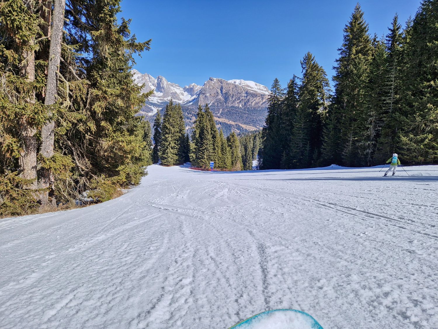 Wide groomed ski run curves through tall evergreen trees with textured snow in the foreground and rugged mountains rising in the distance. A single skier in a bright jacket moves along the right side under a clear blue sky.
Kristine and Emma skied the Bravo ski slope of the Legendary 8 of Val Gardena