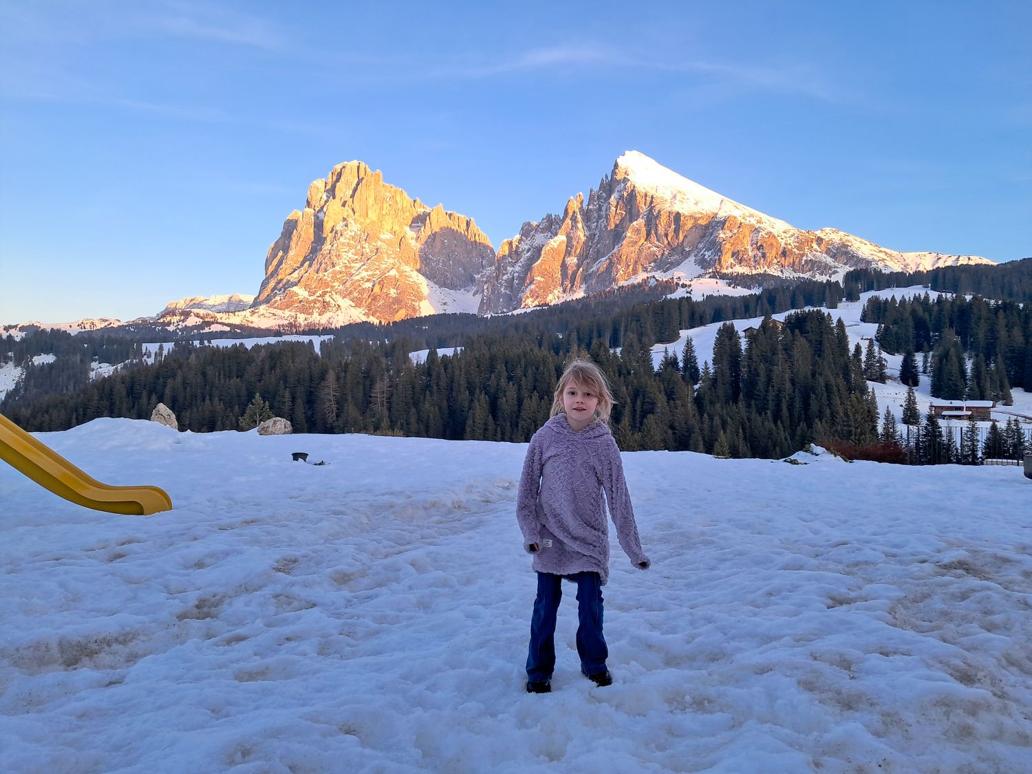 Young child stands on packed snow facing the camera with a forested hillside and dramatic rocky peaks glowing in warm sunset light behind them. A small yellow slide sits off to the left while scattered cabins and trees dot the snowy landscape under a clear sky.