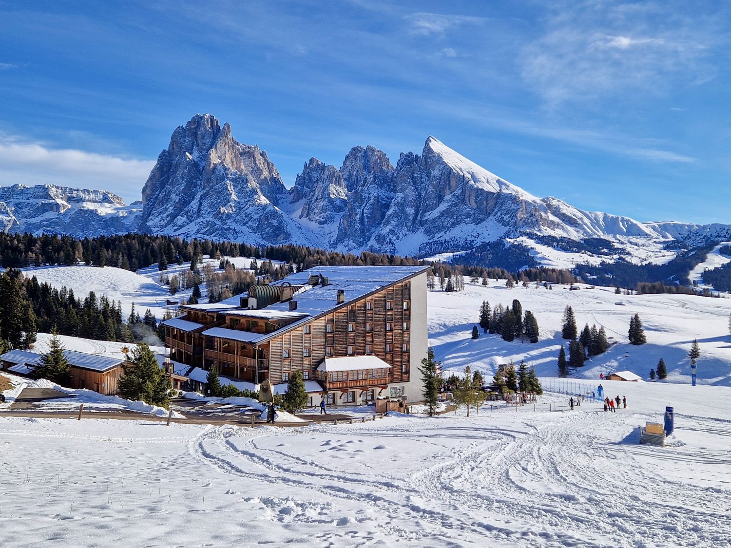A modern alpine lodge sits in a wide snowy meadow with ski tracks leading toward it, surrounded by scattered pine trees. Towering jagged mountain peaks rise sharply in the background under a bright blue winter sky, emphasizing the dramatic high alpine setting.