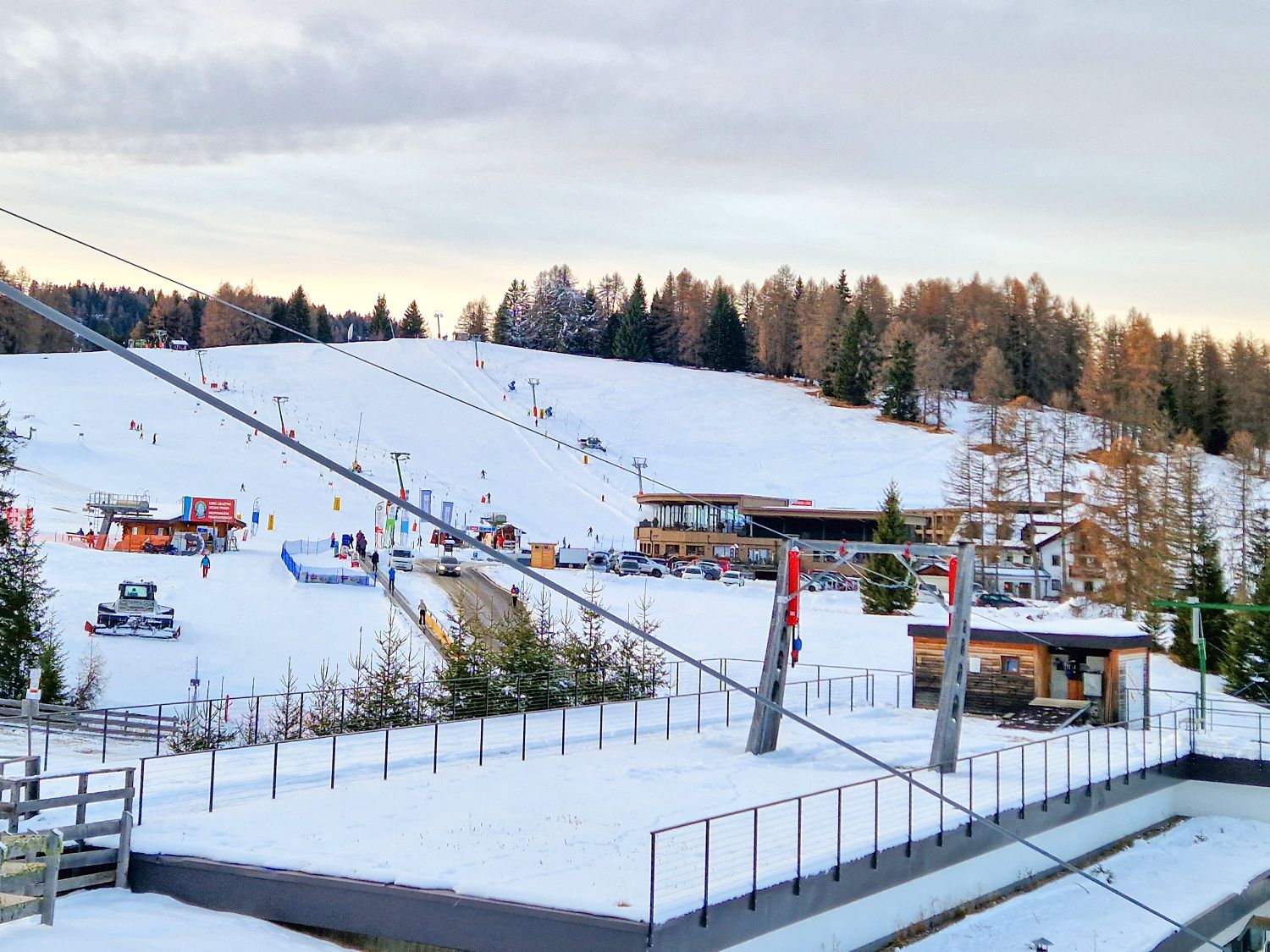 A snowy ski resort with ski lifts, people skiing on the slopes, buildings at the base, parked cars, and tall trees in the background under a partly cloudy sky.
