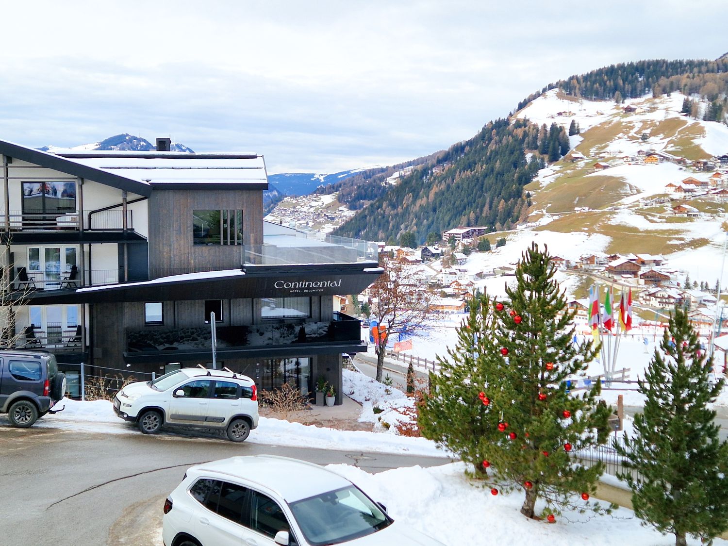 A modern mountain hotel with "Continental" sign stands on a snowy hillside, overlooking a village and mountains. Snow-dusted pine trees with red ornaments and parked cars are in the foreground. Multicolored flags are visible nearby.