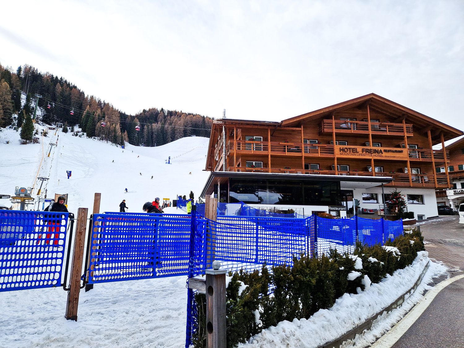 A wooden hotel building sits beside a snowy ski slope with skiers and ski lifts. Blue mesh fences line the path, and evergreen trees cover the hillside in the background under a cloudy sky.