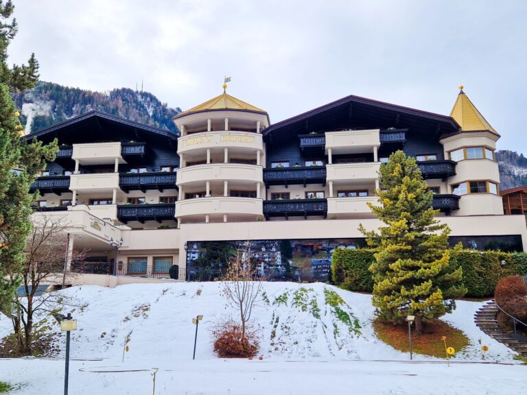 Front view of a luxury mountain hotel with symmetrical balconies and gold roof accents, set against snow covered ground and forested hills. The building appears grand and resort like, emphasizing its scale and alpine architecture.