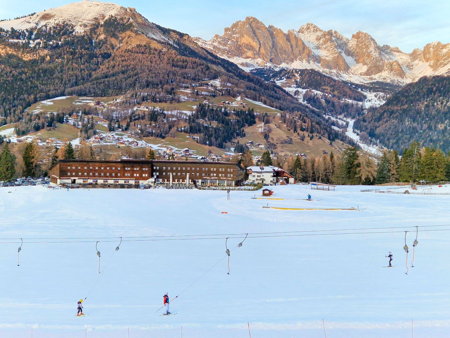 A wide snowy ski slope stretches across the foreground with several skiers using a surface lift. In the background, a long alpine lodge sits at the base of the mountain, with forested hills and rugged, sunlit peaks rising behind it under a clear winter sky.