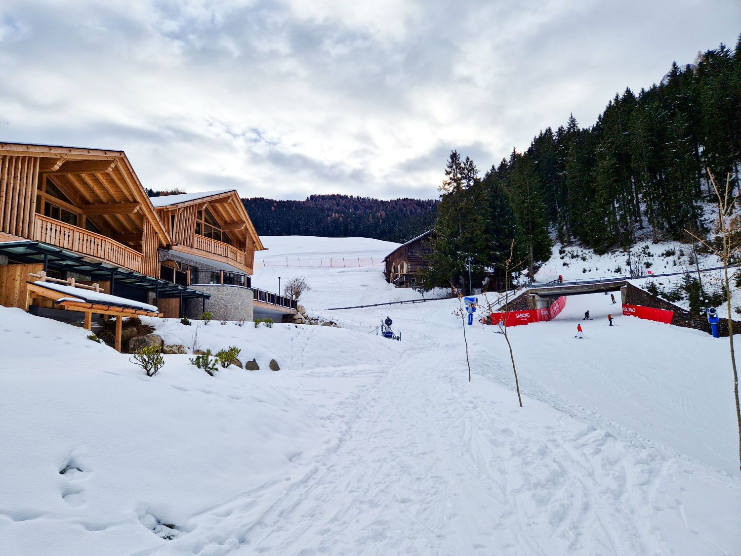 A snowy ski resort with wooden lodges on the left, people skiing on a slope to the right, and pine trees lining the hillside. The sky is cloudy and the area is surrounded by snow-covered ground.