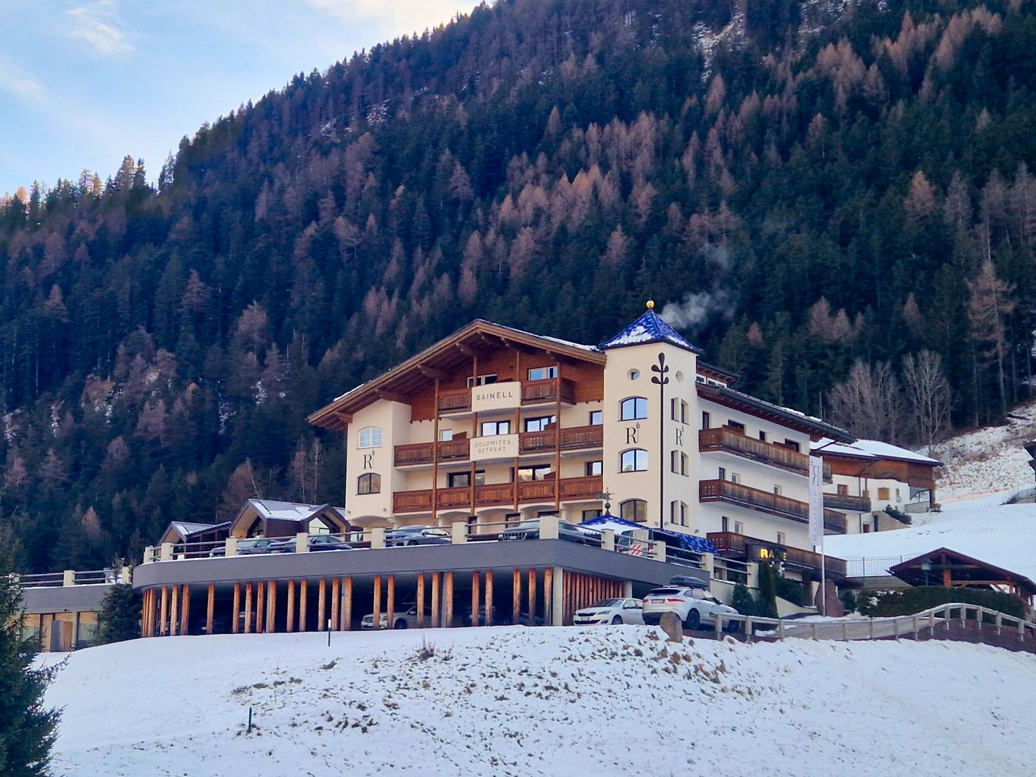 A large alpine hotel sits on a snowy hillside surrounded by dense evergreen forest. The multi story building has wooden balconies and a covered parking area beneath it, with mountains rising behind.