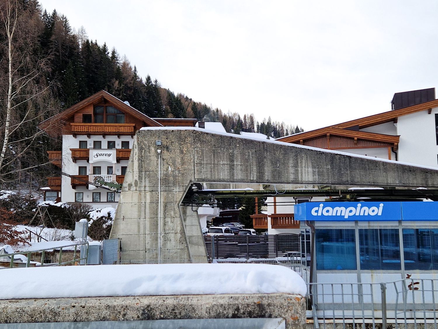 A snowy mountain village with wooden lodges, a concrete structure, and a blue gondola station labeled "ciampinoi" in the foreground, surrounded by evergreen trees.