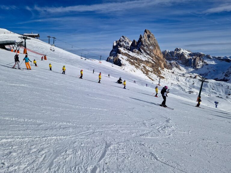 A group of skiers descends a gentle snow covered slope, including several children wearing bright helmets in a ski lesson. Chairlift cables and rocky mountain peaks frame the scene, showing an active ski area on a clear winter day.