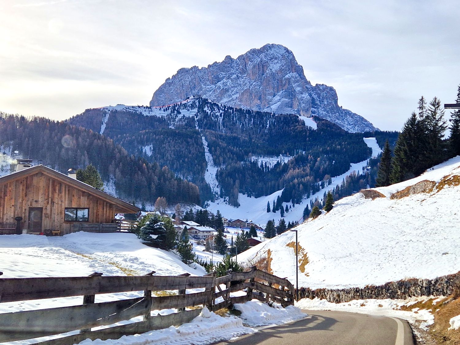 A quiet mountain road curves through snow covered hills toward a small alpine village, with a wooden chalet in the foreground. A dramatic rocky mountain rises in the background above forested slopes and visible ski runs, creating a peaceful winter landscape.