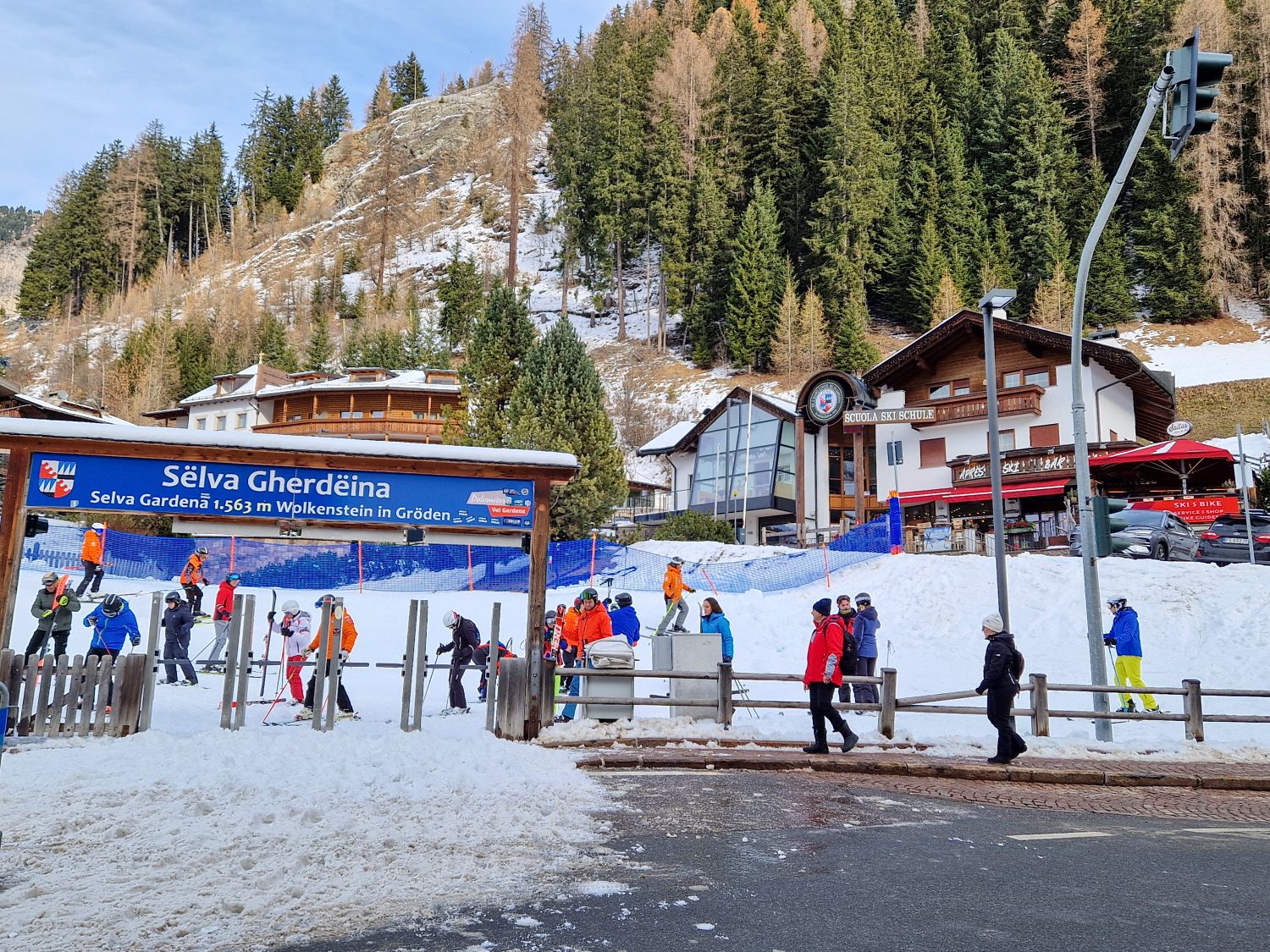 A busy ski school area in Selva Gardena shows skiers lining up beneath a large sign reading Selva Gherdeina Selva Gardena 1,563 m Wolkenstein in Gröden. People in colorful ski gear gather near beginner slopes and fencing, with alpine buildings and forested mountains rising behind the village setting.