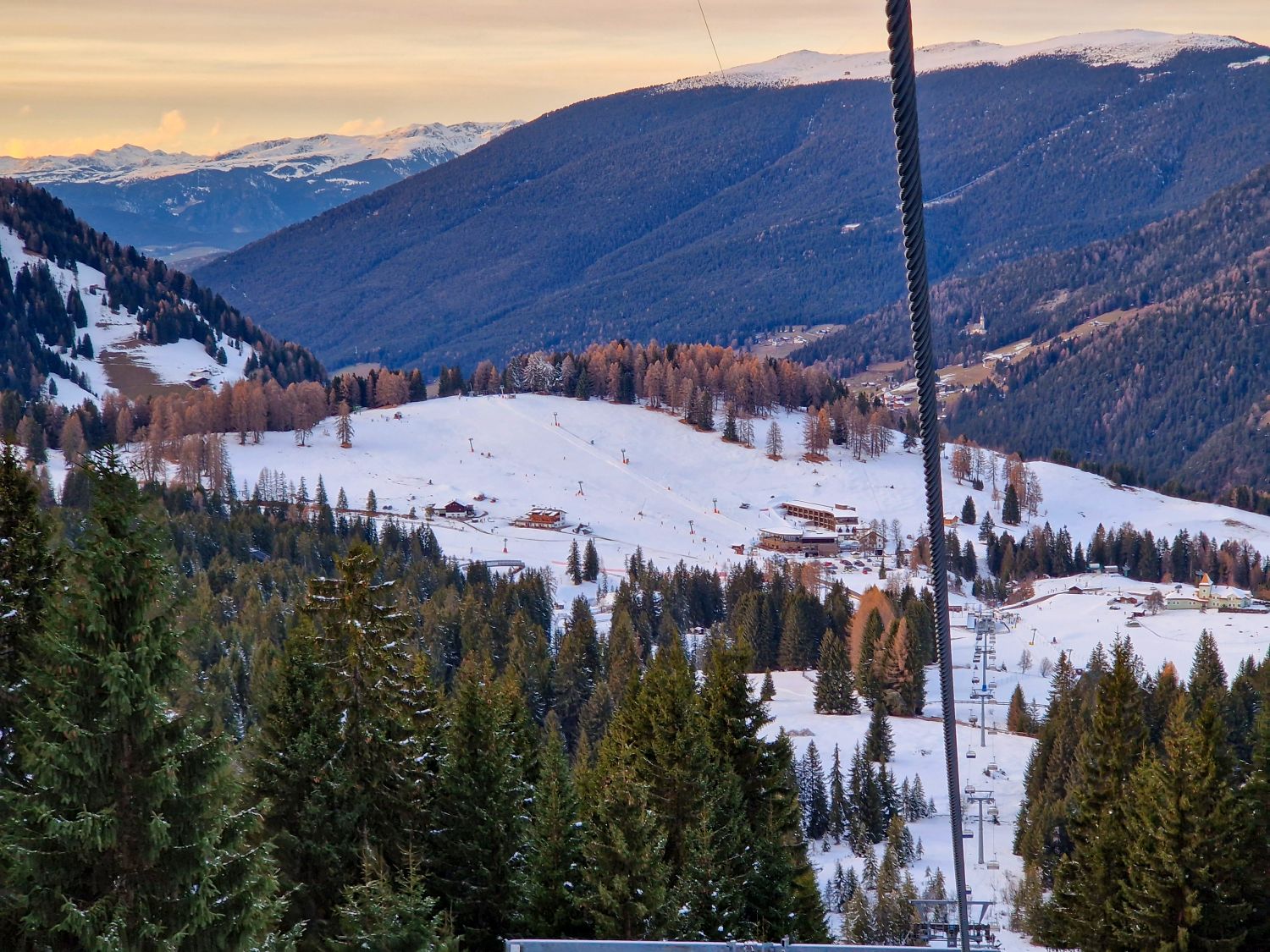 A ski area sits in a snowy valley with chairlift lines descending through evergreen trees toward lodges and open beginner slopes. Forested mountains rise in layered ridges beyond the resort, with warm late day light touching the distant peaks.