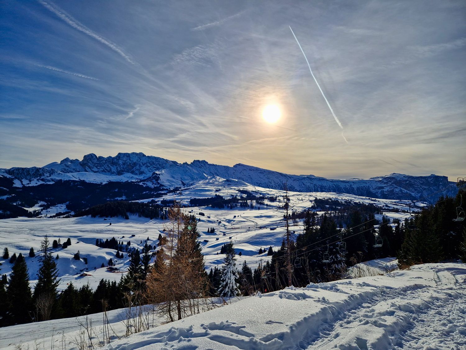 A wide panoramic view shows rolling snow covered meadows and groomed ski terrain stretching across a high alpine plateau. The low winter sun glows through streaked clouds above distant mountain ridgelines, with dark pine forests framing the foreground snow.