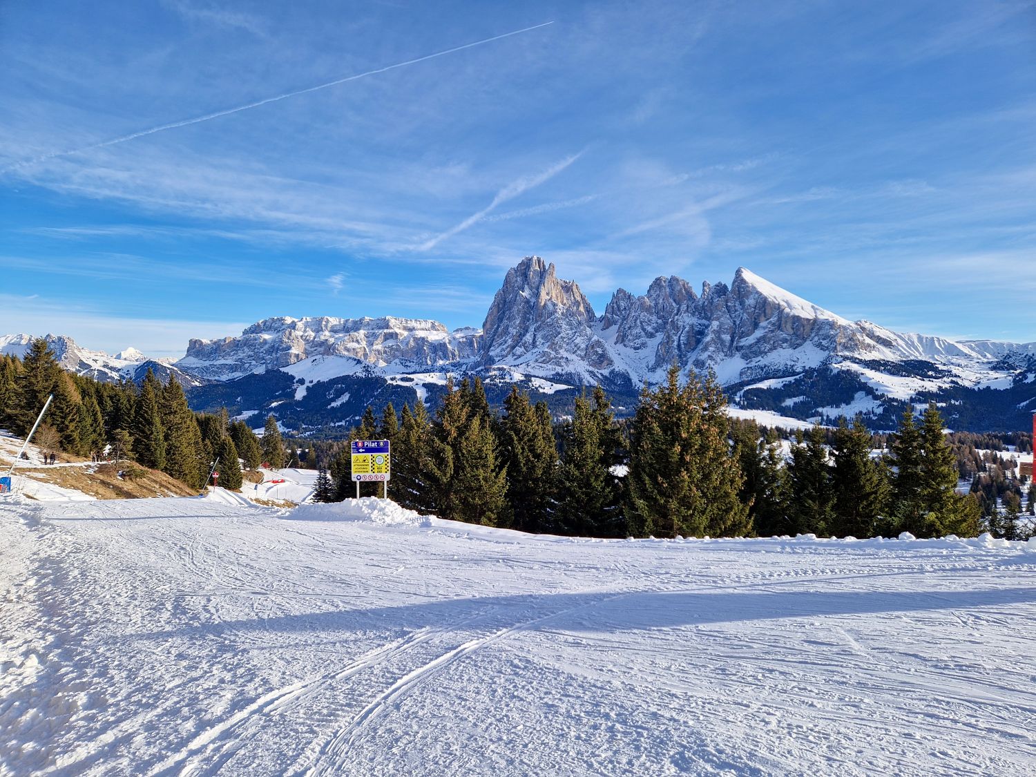 A groomed ski trail opens onto a panoramic view of jagged snow covered mountain peaks under a bright blue sky. Evergreen trees line the slope, with ski tracks in the foreground and a trail sign marking the route through the alpine landscape.