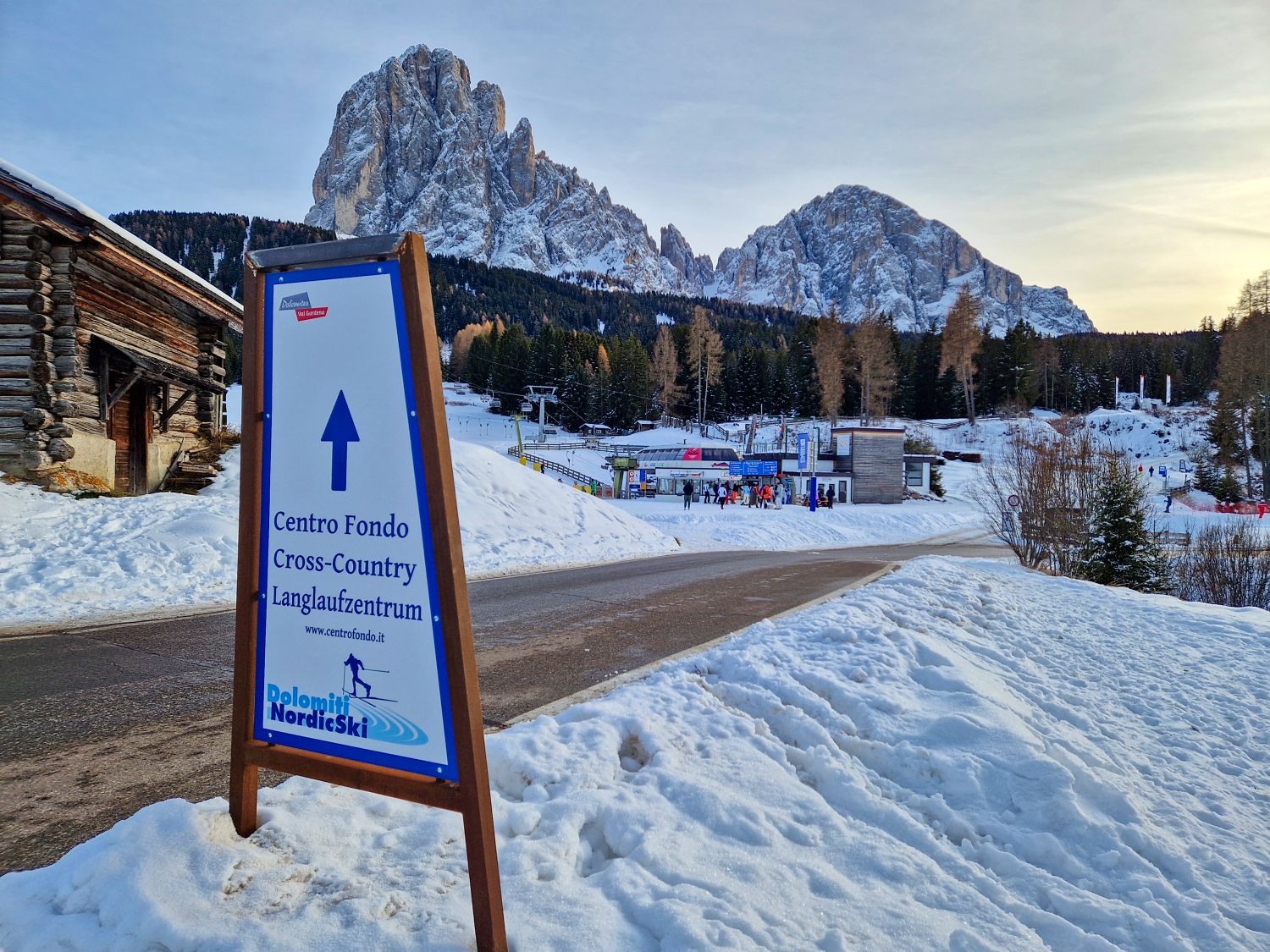 A roadside sign in the snow points toward Centro Fondo Cross Country Langlaufzentrum with an arrow and Dolomiti NordicSki logo. The sign stands beside a cleared road leading to a ski area, with wooden buildings, people in the distance, and dramatic snow covered Dolomite peaks rising behind evergreen trees.