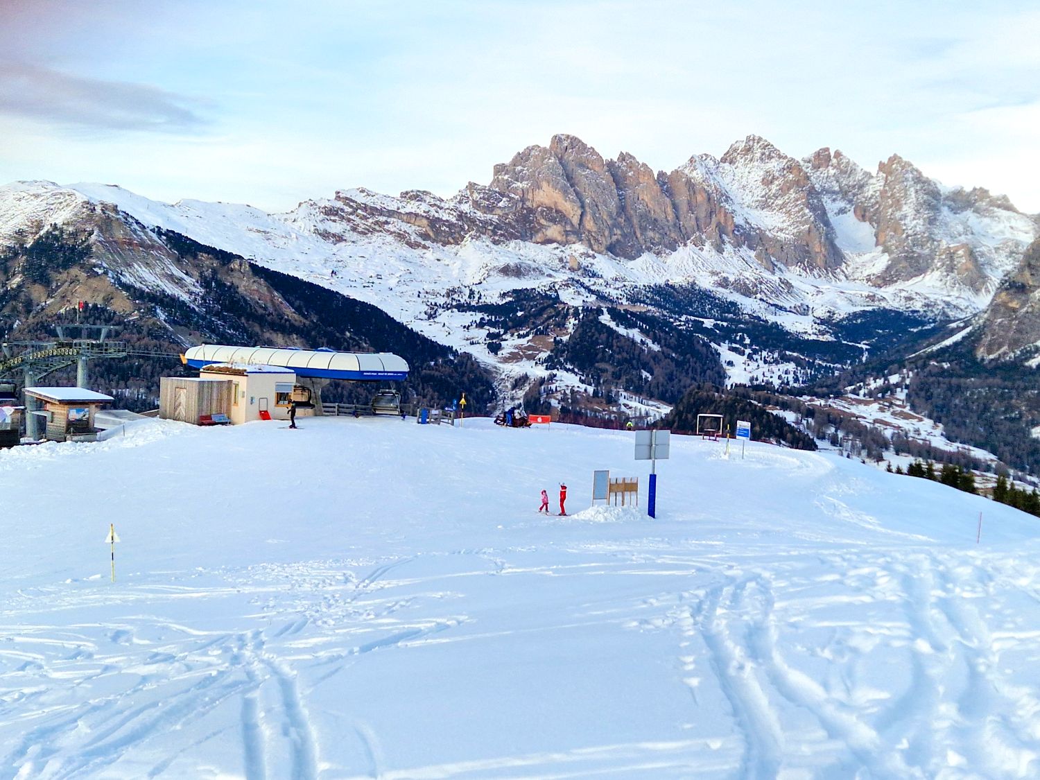 A wide snowy plateau at the top of a ski area features a lift station and open beginner terrain. Two small skiers stand near trail signs while dramatic rocky mountain peaks and layered alpine valleys stretch across the background under a pale winter sky.