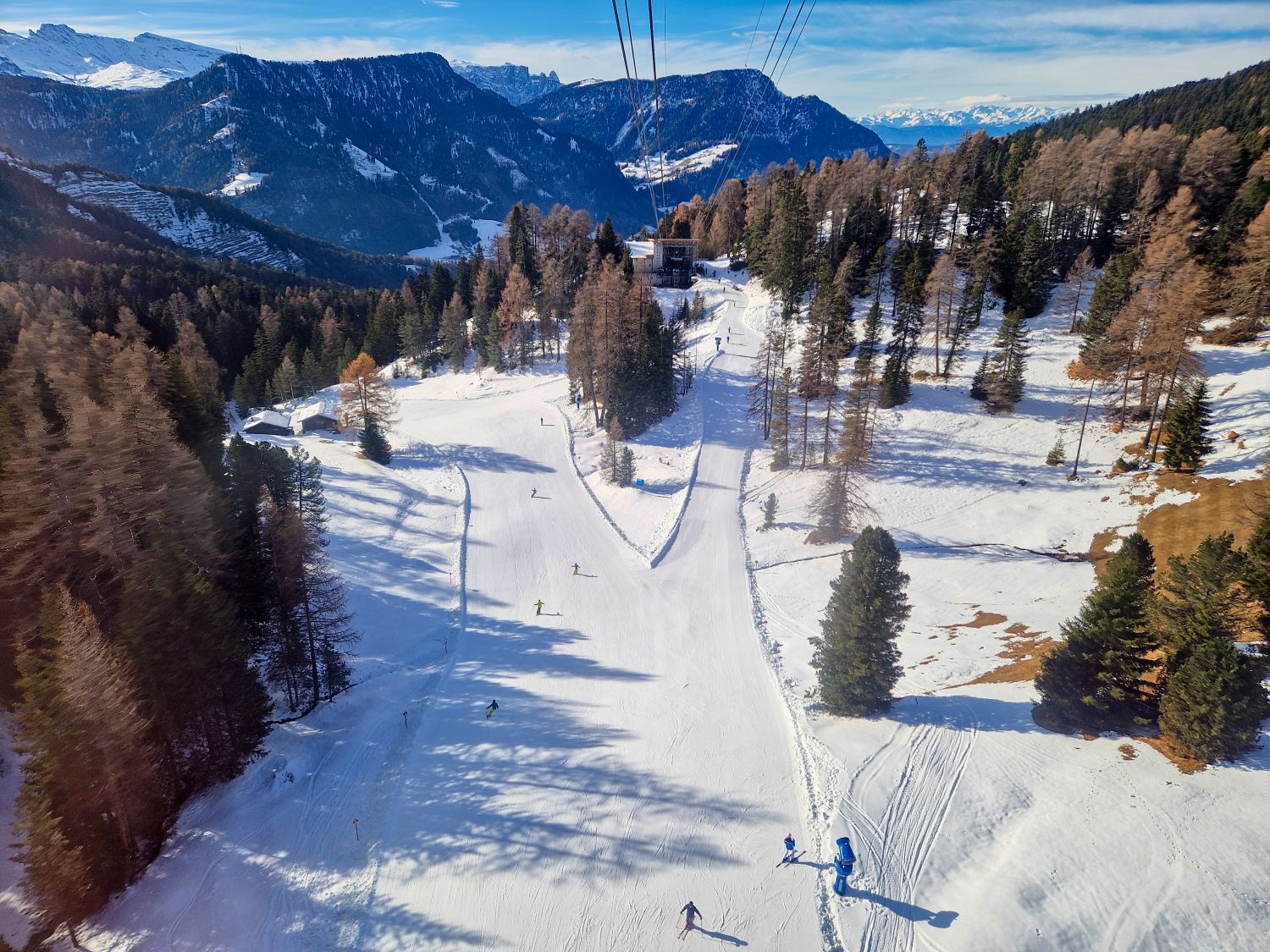 An aerial view looks down over a groomed ski run winding through snow covered forest, with several skiers descending the slope below lift cables. Rolling alpine hills and distant mountain ridges stretch across the background under a clear blue winter sky.