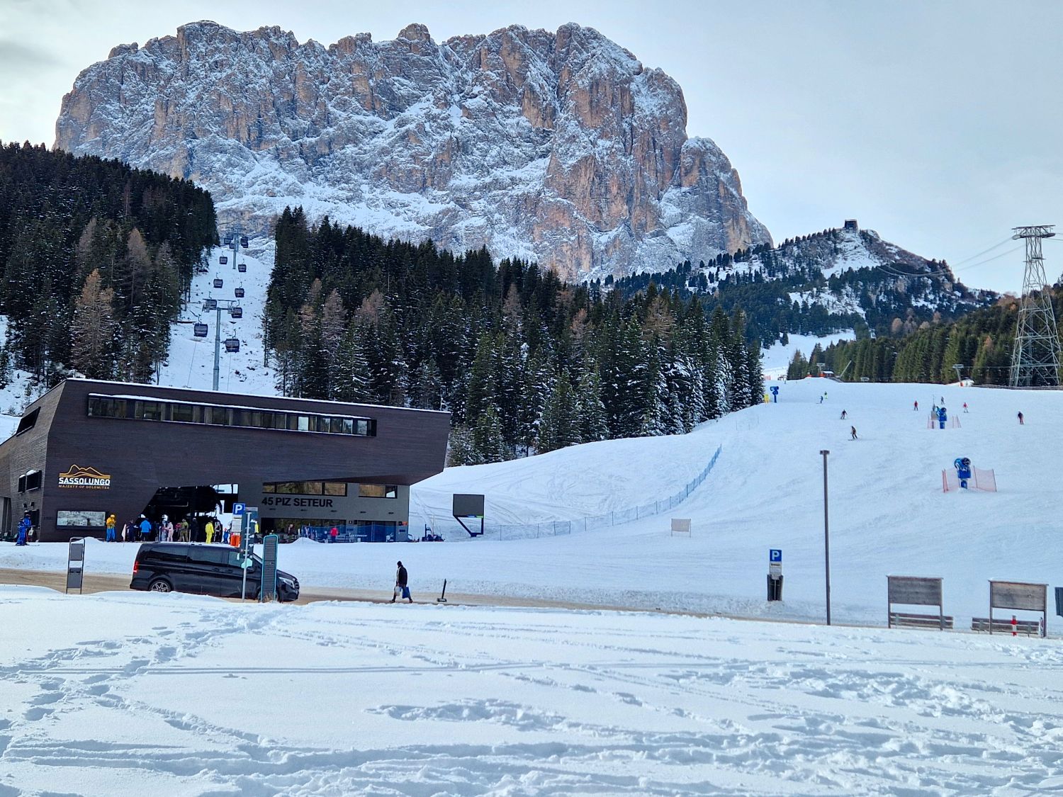 A modern ski lift station sits at the base of a wide snow covered slope, with skiers ascending and descending nearby. Dense evergreen forest lines the run, while a massive rocky mountain rises behind the lift, dominating the winter alpine landscape.