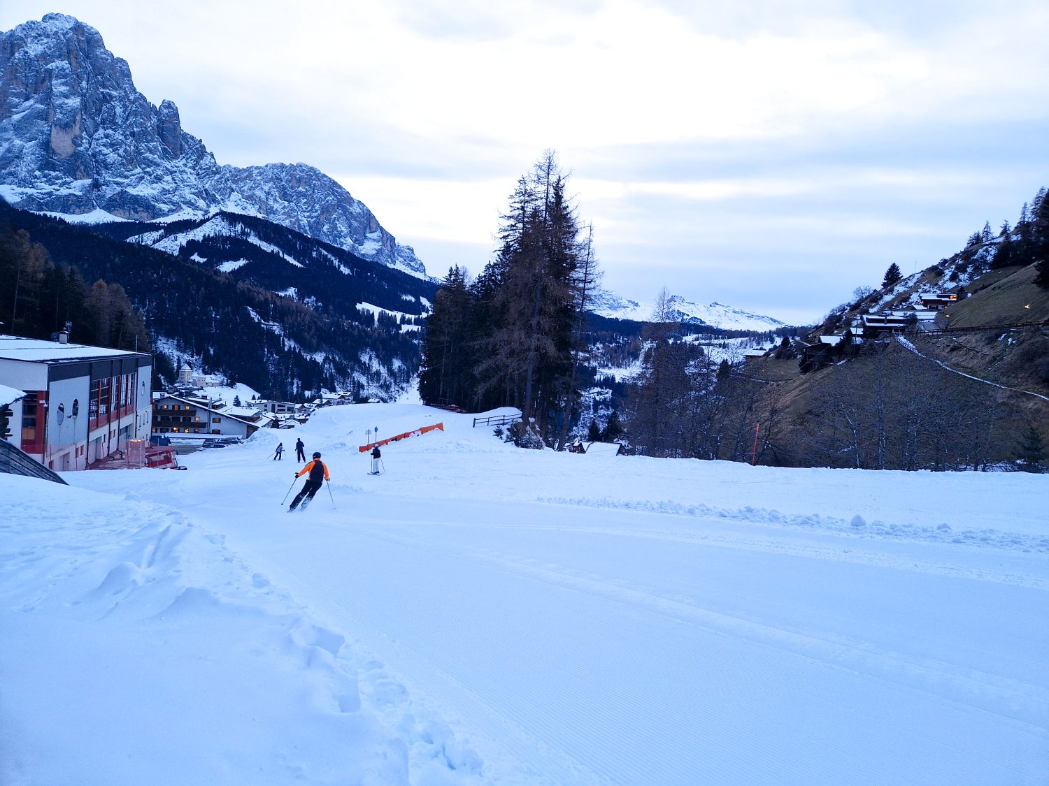 A groomed ski run curves downhill toward a mountain village, with a skier carving across the snow in the foreground. Forested slopes and dramatic rocky peaks rise in the background, framing a quiet alpine resort scene on a winter day.