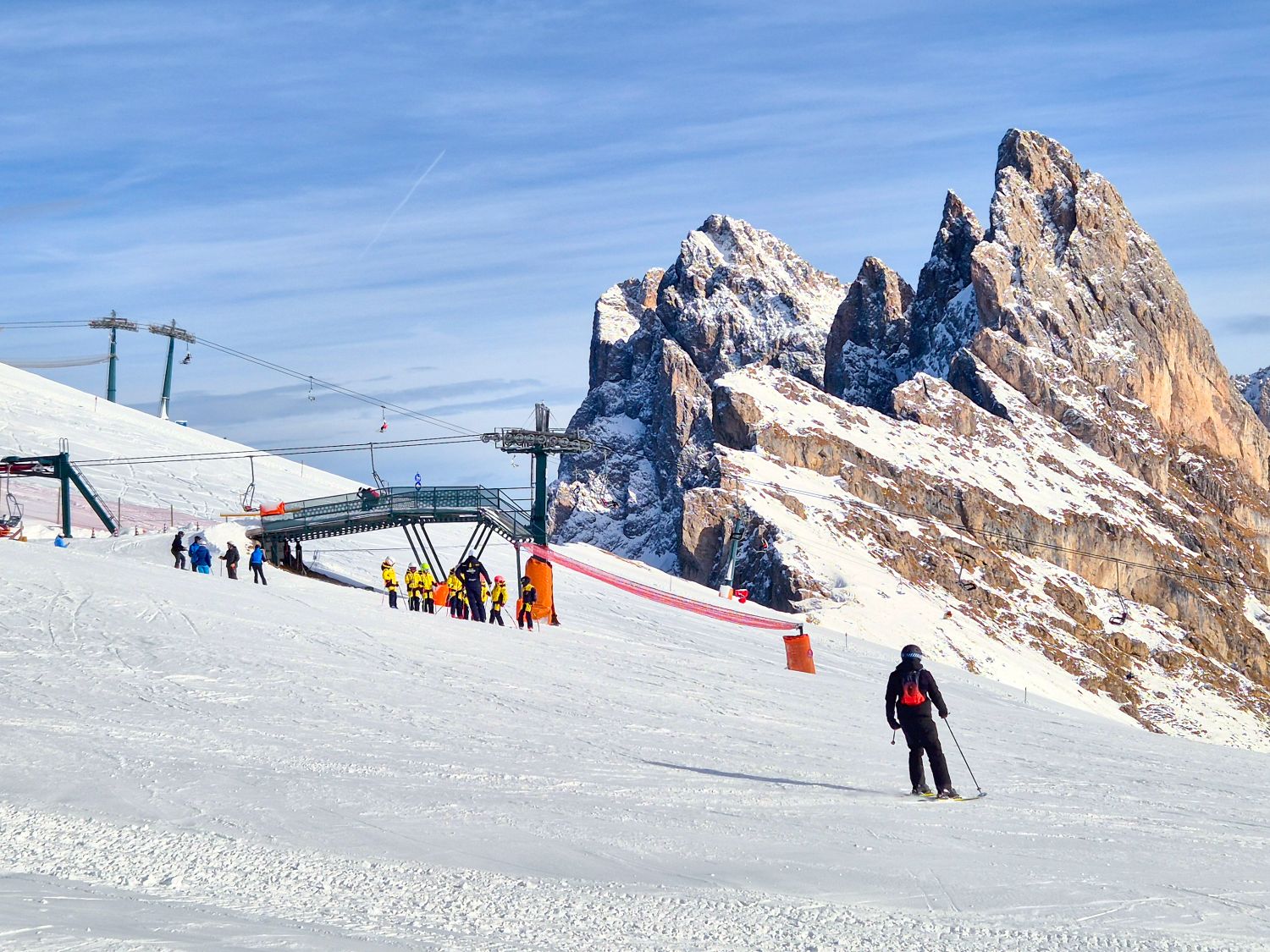 A ski lift station sits on a snowy slope with a small group of skiers gathered nearby while one skier glides downhill in the foreground. Jagged rocky mountain peaks rise sharply behind the lift under a clear blue winter sky, emphasizing the dramatic high alpine setting.