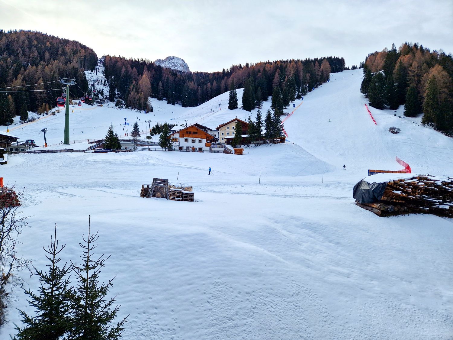 A wide snowy ski valley opens around a cluster of alpine chalets at the base of multiple groomed slopes. Chairlifts climb the hillsides through evergreen forests while a few skiers cross the open snow under a calm winter sky.