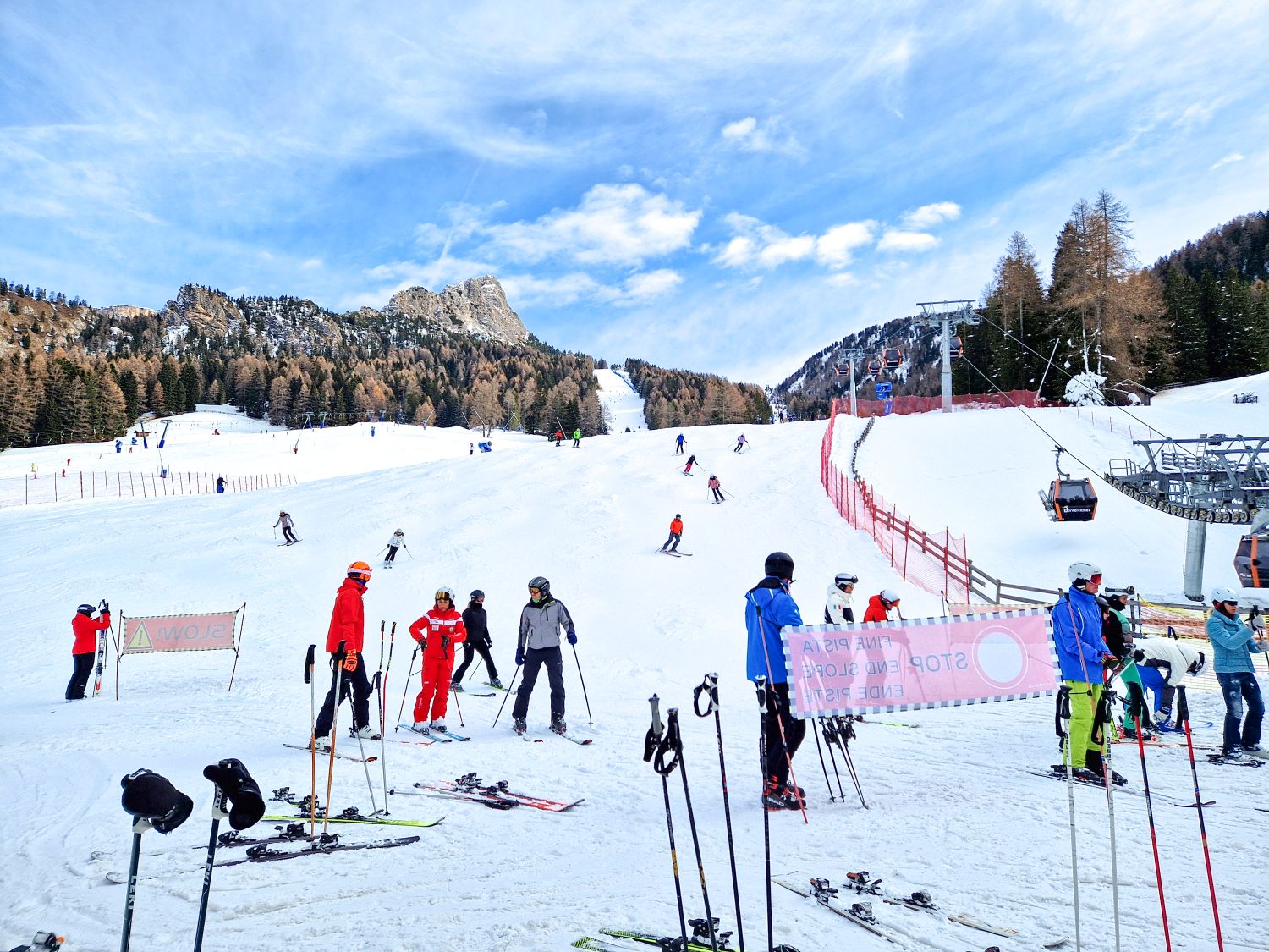 A wide ski slope is busy with skiers gathered at the bottom while others descend from above under a bright blue sky. Skis and poles rest in the snow near warning signs, with chairlifts, safety fencing, and forested mountains framing an active winter resort scene.