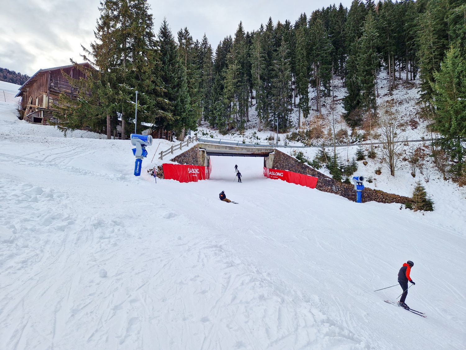 A snow covered ski slope passes beneath a small road bridge, with skiers descending through a narrow underpass lined with red safety padding. Snowmaking machines stand on either side of the run, and evergreen trees and a wooden chalet frame the winter mountain setting.