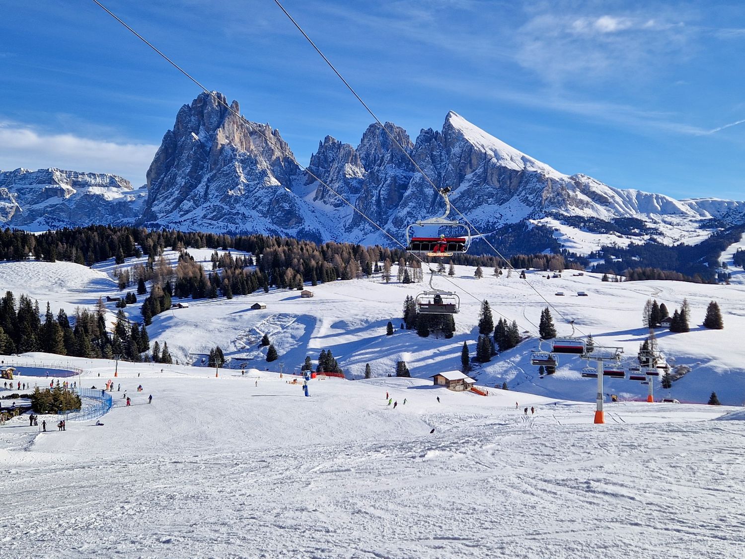 A wide ski slope stretches downhill beneath chairlift cables, with skiers scattered across the snow. Rolling white meadows dotted with evergreen trees lead to dramatic jagged mountain peaks rising sharply in the background under a clear blue winter sky.